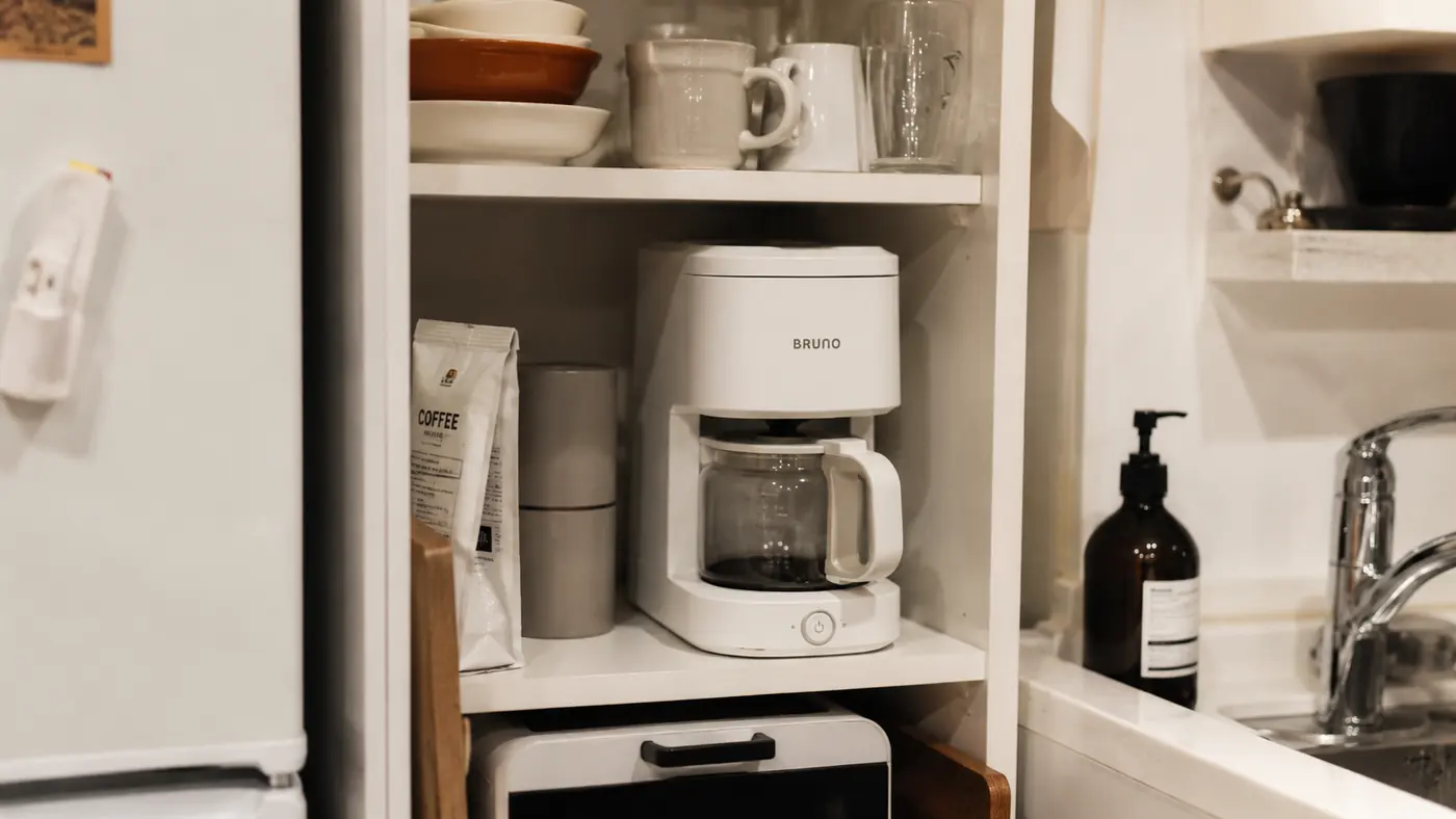 Keurig K-Mini Mate stored on a narrow shelf between a cutting board and a stack of mugs in a small apartment kitchen