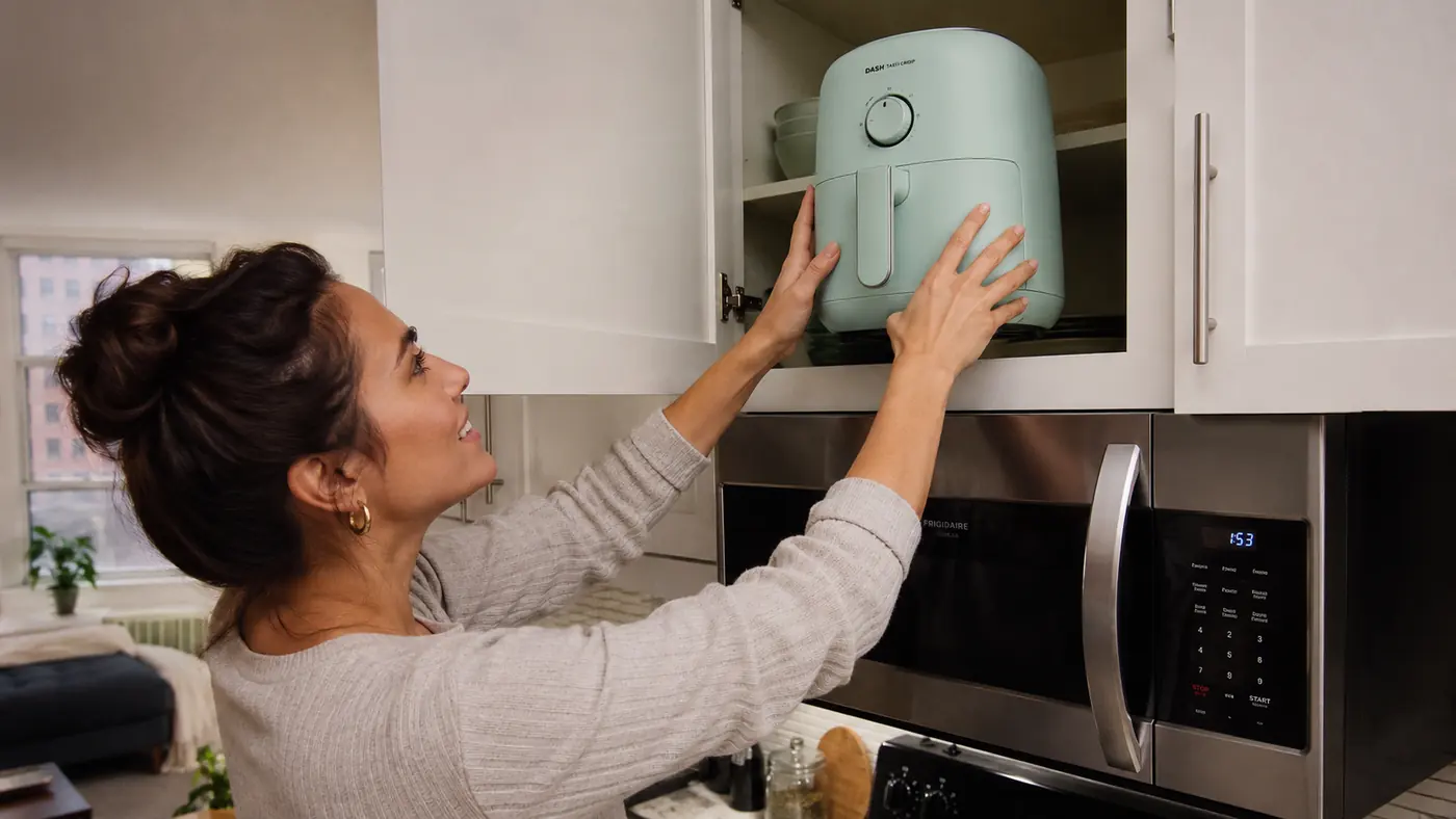 Hand lifting a small, light air fryer into an overhead kitchen cabinet with one hand