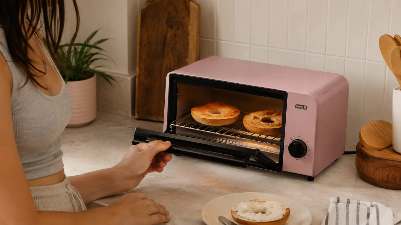 Hand opening the door of the Dash Mini Toaster Oven to check on two toasting bagel halves