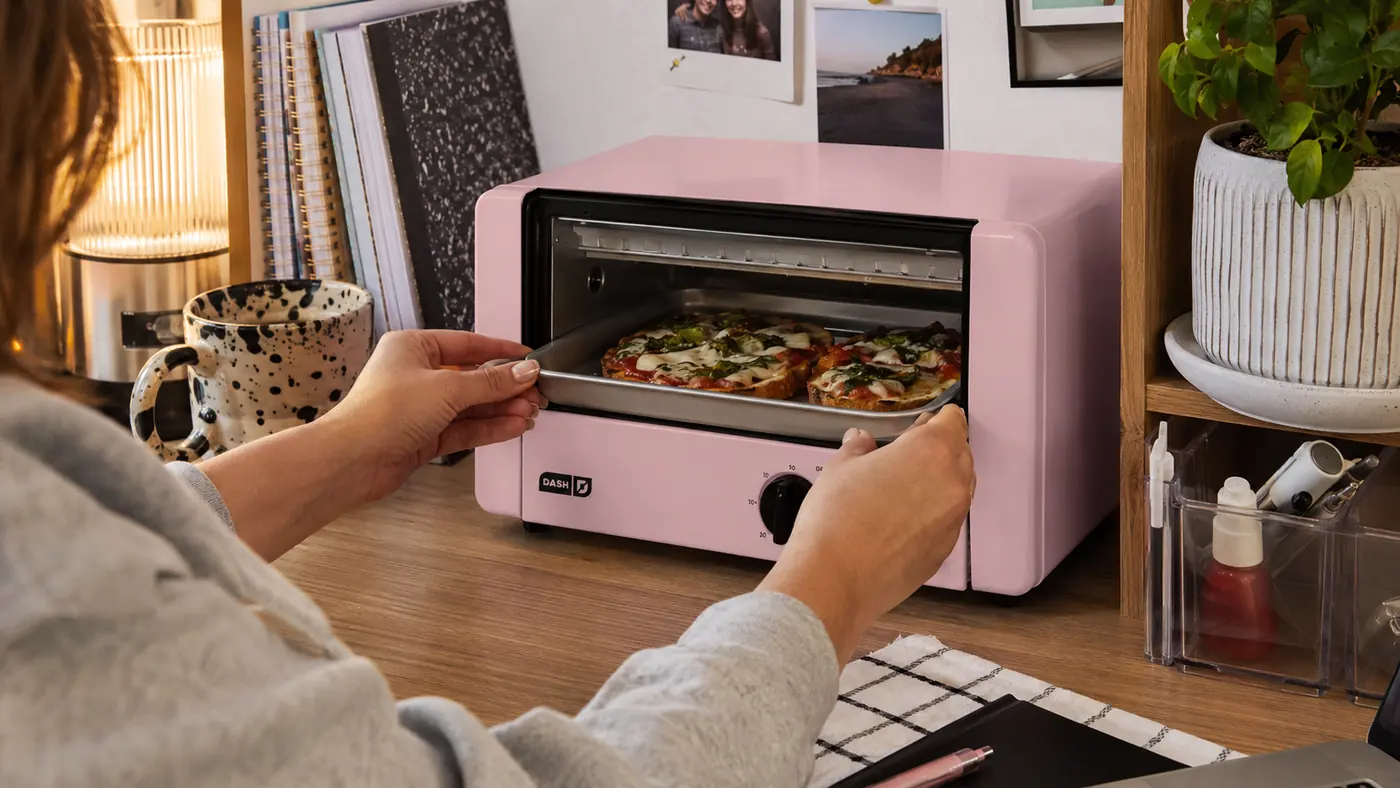 Hand sliding a small metal tray of food into the Dash Mini Toaster Oven on a dorm desk