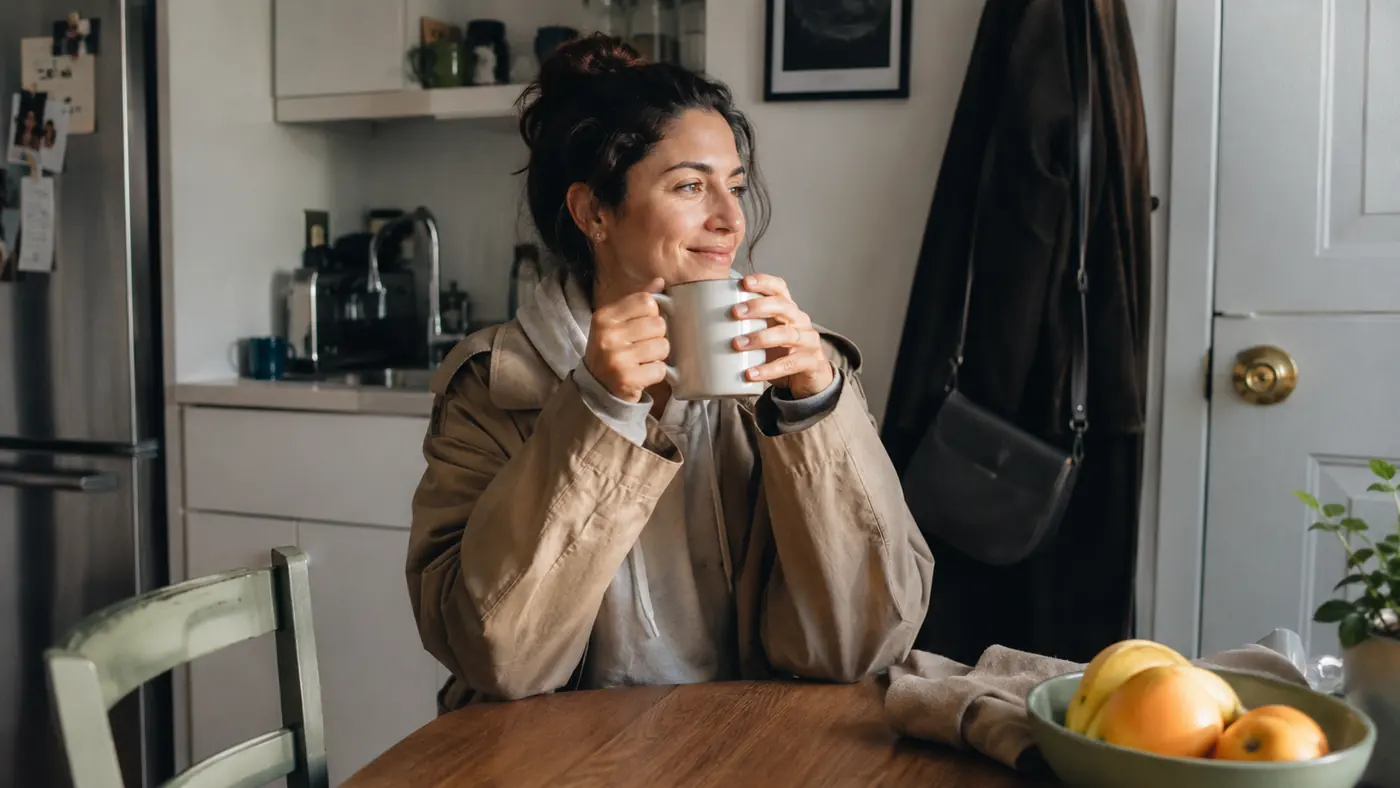 Bethany holding a mug of coffee at her small kitchen table, coat already on, ready to leave for work
